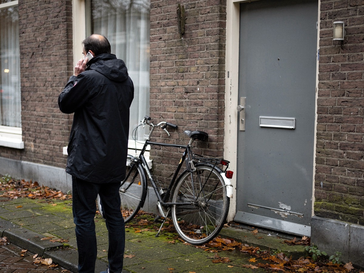 Person standing on the pavement outside a Dutch terraced house, phone to their ear, asking a locksmith for the full price
